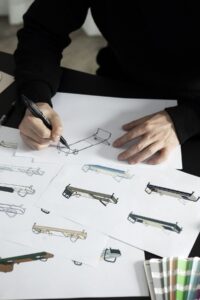 Designer drawing a sketch of a Pilates reformer or fitness equipment design on paper, surrounded by color swatches and other drawings.