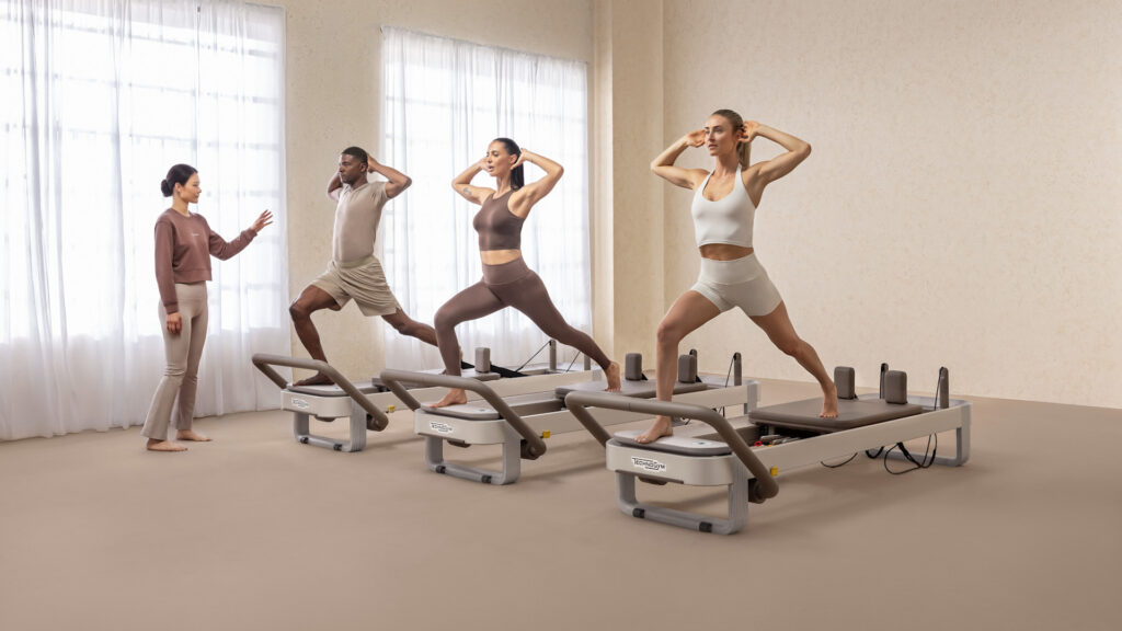 Pilates instructor guiding a diverse group of three students performing a standing lunge exercise on TechnoGym reformers.
