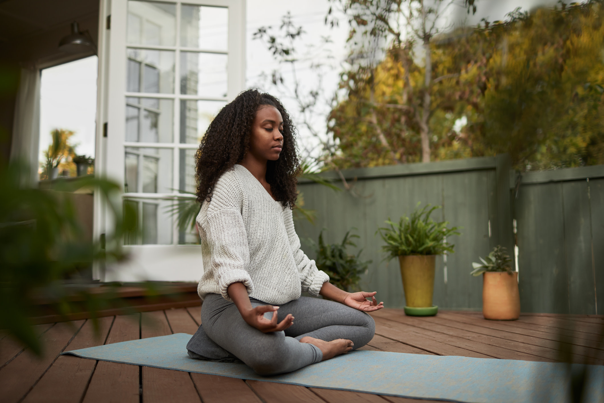 A woman sits cross-legged on a yoga mat outdoors, meditating with her eyes closed and hands in a mudra position.