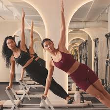 Two people performing a side plank exercise on Pilates reformer machines in a modern studio.